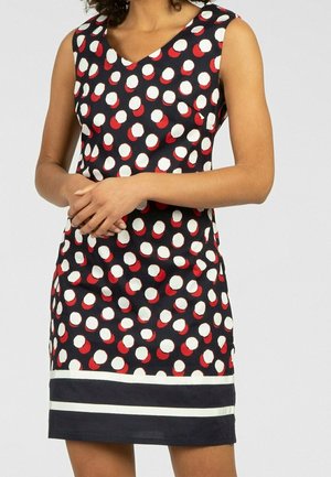 Woman wearing sleeveless black dress with white and red polka dots and two white horizontal stripes near the hem, hands clasped.