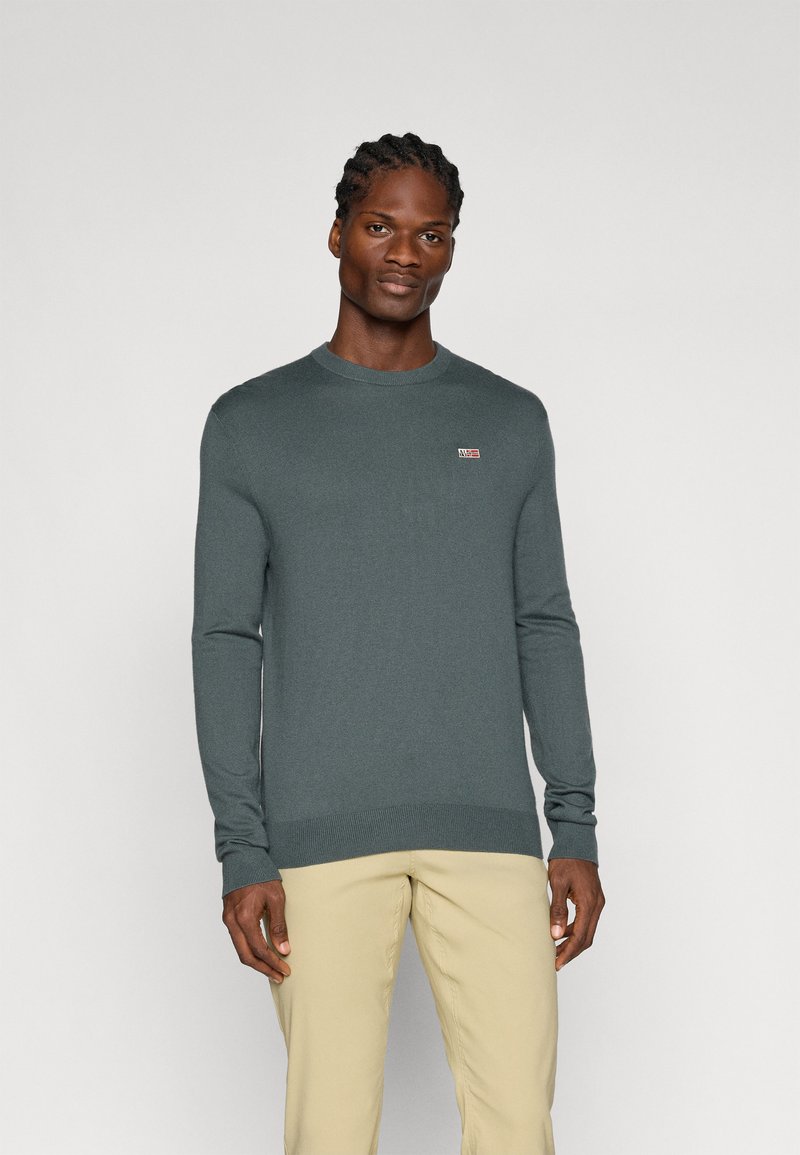 Pull en tricot vert foncé à col rond, à manches longues, avec des poignets et un ourlet en côtes. Présente un petit patch logo sur la poitrine.