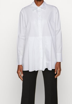 Personne portant une chemise blanche à manches longues avec col et un pantalon noir plissé, debout devant un fond uni.