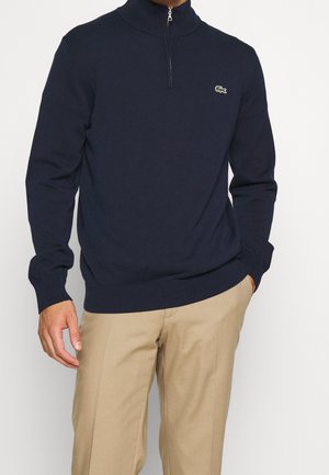 Mann trägt einen marineblauen Half-Zip-Pullover mit Lacoste-Logo und beige Hosen, steht vor einem schlichten hellen Hintergrund.