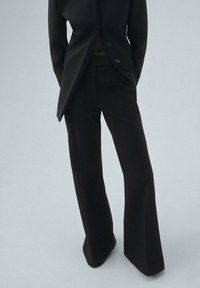 Black tailored suit jacket with a pointed hem and two buttons, paired with black wide-leg trousers, both made from a smooth fabric.
