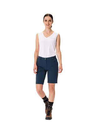 Marineblauwe shorts met een getailleerde pasvorm, knoopsluiting en ritszakken, gecombineerd met een witte mouwloze top en bruine wandelschoenen.