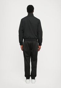 Black windbreaker with a high collar, elastic waistband, and matching black trousers. White sneakers complete the outfit. Smooth texture.