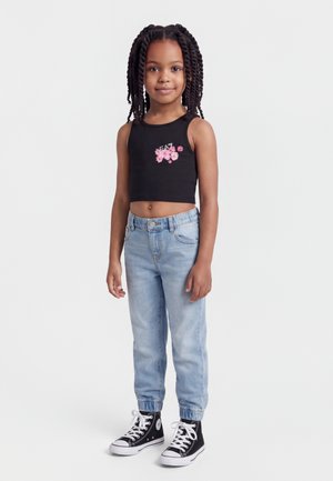 Fiatal lány copfos hajjal, fekete crop topot visel rózsaszín virágokkal, világoskék farmert és fekete magas szárú tornacipőt, állva.