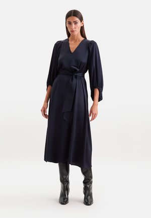 Frau trägt ein marineblaues Midikleid mit langen Puffärmeln, V-Ausschnitt, gebundenem Taillengürtel und schwarzen kniehohen Stiefeln und steht vor weißem Hintergrund.