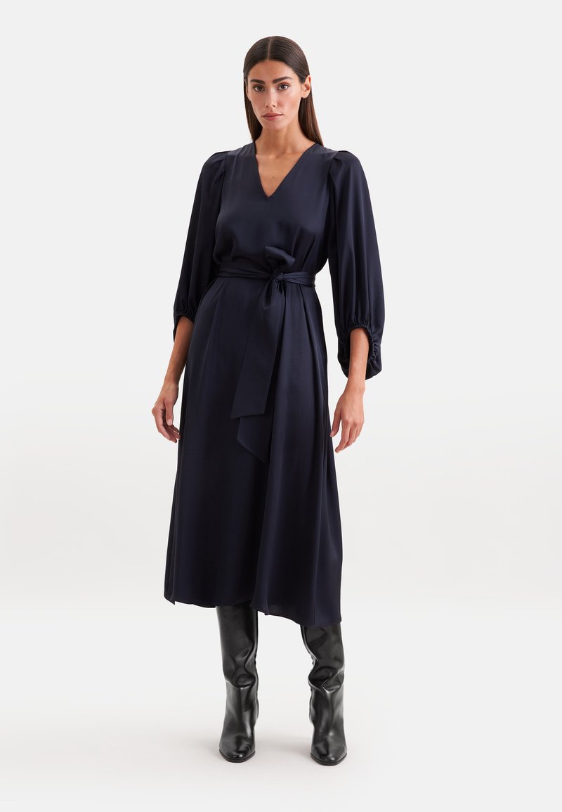 Femme portant une robe midi bleu marine avec de longues manches bouffantes, un col en V, une ceinture nouée à la taille et des bottes noires montantes, debout sur un fond blanc.