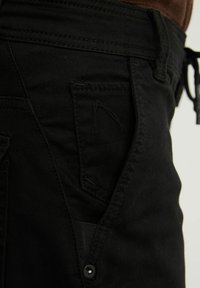 Gros plan sur une poche de pantalon noire avec coutures visibles, un petit rabat et un détail bouton sur le côté près de la ceinture.
