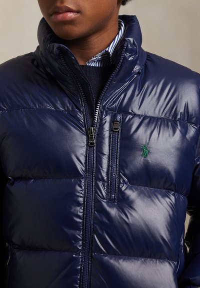 Jeune personne portant une doudoune bleu marine brillante avec un logo vert brodé visible sur la poitrine et un col de chemise rayé.