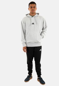 Felpa grigia con tasca frontale e logo Adidas nero, abbinata a pantaloni jogger neri con un logo bianco, completata con scarpe scure.