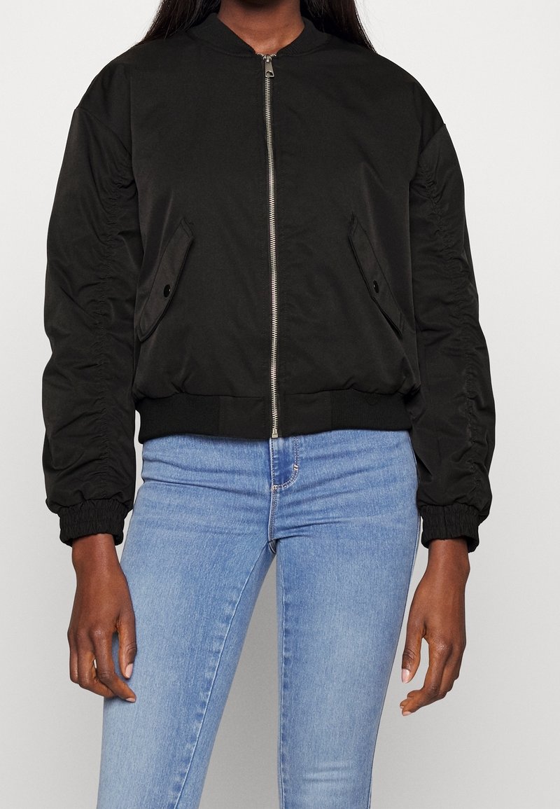 Femme portant une veste bomber noire zippée avec des poches avant et des manches froncées, associée à un jean en denim bleu clair.