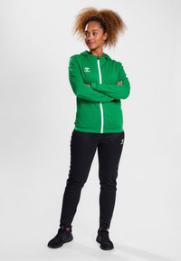 Groene zip-up hoodie met witte accenten, gecombineerd met zwarte sportbroek. Soepel materiaal met een relaxte pasvorm, ideaal voor sportkleding.
