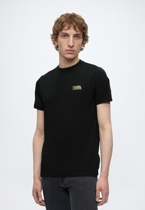 Giovane uomo con capelli ricci che indossa una T-shirt nera Karl Lagerfeld e jeans scuri, in piedi davanti a uno sfondo bianco semplice.