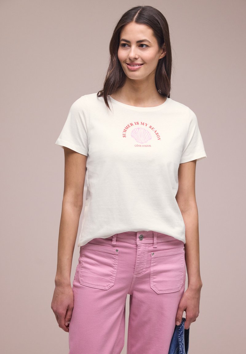 Jeune femme portant un T-shirt blanc avec le texte « L'été est ma saison » et un pantalon rose, tenant une veste en jean bleu dans sa main droite.