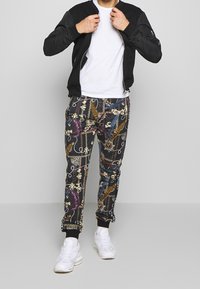 Chaqueta bomber negra sobre una camiseta blanca, combinada con pantalones estampados que tienen detalles coloridos. Zapatillas blancas completan el conjunto.