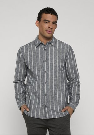 Mann trägt ein graues, geknöpftes Hemd mit weißen vertikalen Streifen, Hände in den Taschen, steht vor einem einfachen hellen Hintergrund.