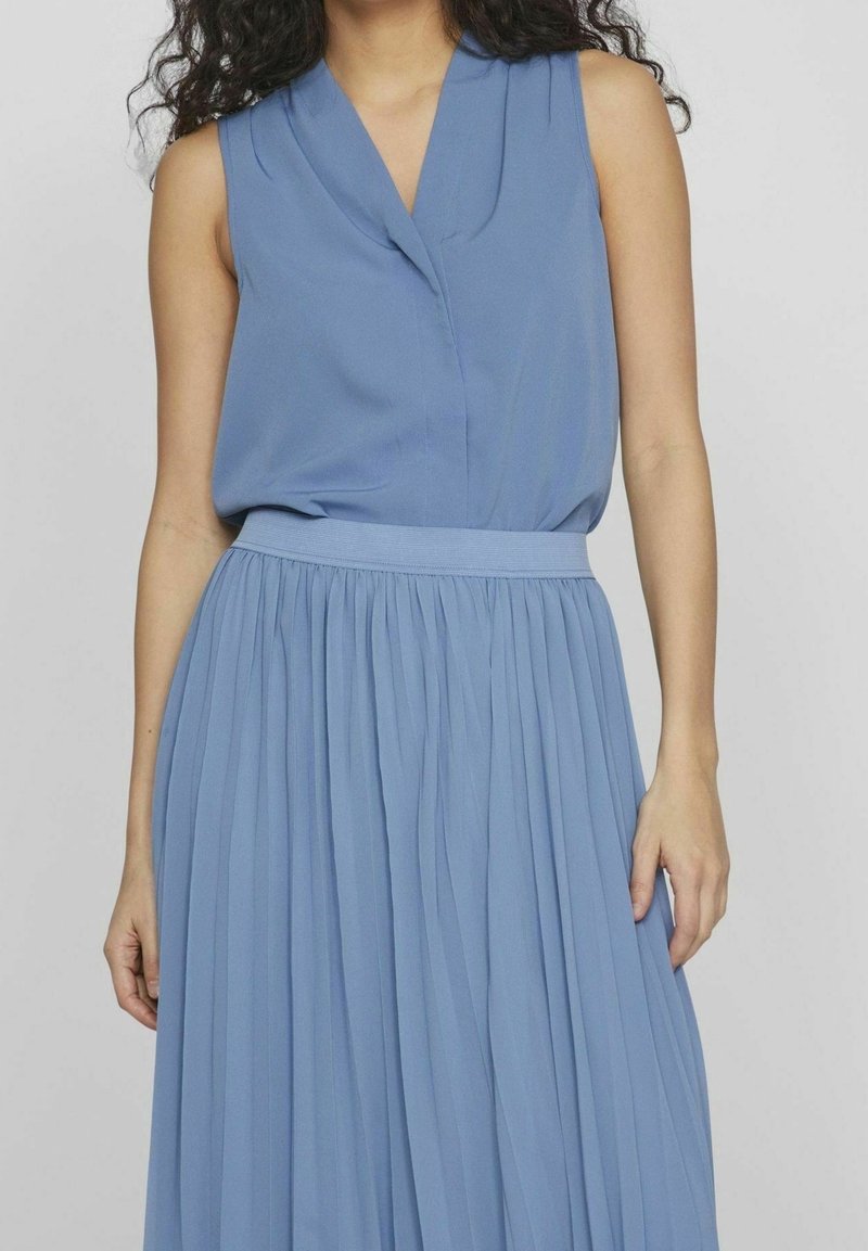 Femme portant une robe plissée bleu clair sans manches avec une taille cintrée sur un fond uni.