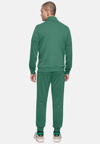 Chándal verde hecho de tela suave. Cuenta con una chaqueta con cremallera y cuello, y pantalones a juego con puños elásticos. Acentuado con calzado con logo.