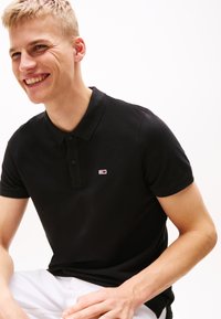 Črna polo majica iz bombaža, z teksturirano tkanino, klasičnim ovratnikom, dvema gumboma in majhnim logotipom s rdečimi in modrimi akcenti.