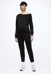 Svart bomulls off-shoulder sweatshirt kombinerad med matchande avsmalnande byxor, bärs med en svart keps och vita sportstrumpor.