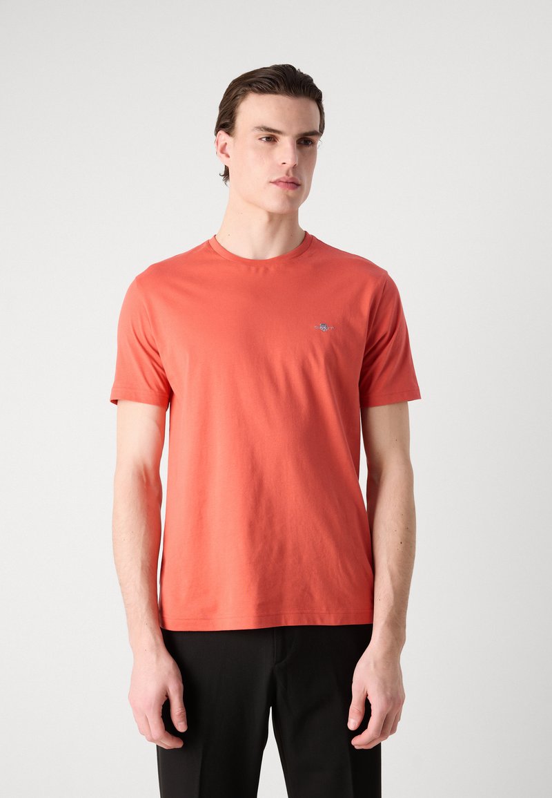 Kurzärmliges T-Shirt aus korallenroter Baumwolle mit Rundhalsausschnitt und kleinem gestickten Logo auf der Brust. Einfaches, schnörkelloses Design.