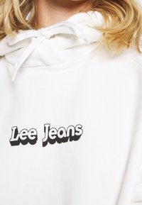 Sudadera blanca con capucha y cordón, que presenta un destacado logo negro de "Lee Jeans". Textura suave, corte relajado y sin patrones ni acentos adicionales.