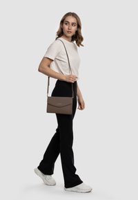 Borsa a tracolla marrone con texture, chiusura a pattina e tracolla lunga, abbinata a una camicia chiara e pantaloni neri. Accessori dal design semplice.