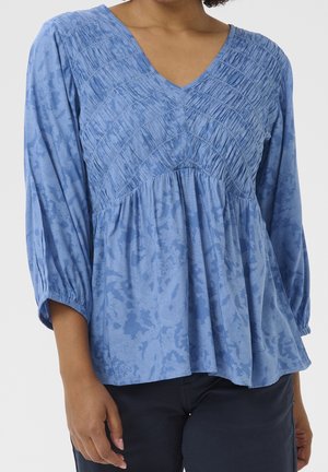 Žena v modré blůze s výstřihem do V, se shromážděným žebrováním na hrudi a jemným květinovým vzorem, v kombinaci s tmavými kalhotami.