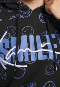 Černá mikina s kapucí, na které jsou modré smajlíky, světle modrá bloková písmena a bílé kurzívou vyšívané písmo přes hrudník, kterou nosí osoba upravující kapuci.