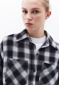 Svart och grå rutigt knapp-up skjorta med två framfickor, strukturerad krage, och ett vitt crew neck lager under.