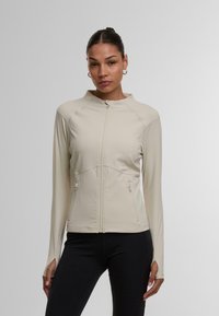 Lys beige atletisk jakke med høj krave, fuld lynlås og to sidelommer. Lavet af glat, strækbart materiale, tætsiddende design, lange ærmer.