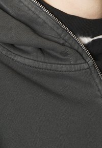 Närbild av en delvis dragkedjad mörkgrå hoodie över en svart skjorta med en vit rand nära halslinningen.