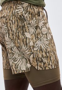 Σύντομο σορτς με καμουφλάζ σχέδιο και λουλουδάτα μοτίβα φορεμένα πάνω από μονόχρωμα ελιτί χρώματος σορτς συμπίεσης σε άτομο με σκουρόχρωμο δέρμα.