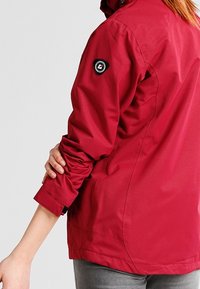 Personne portant une veste rouge avec une manche retroussée tenant l'autre bras, montrant un petit écusson circulaire noir avec logo sur le haut du bras.