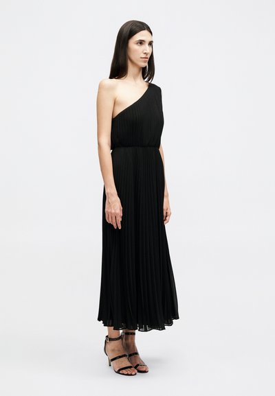 Γυναίκα με μαύρο μίντι φόρεμα με ένα ώμο και πτυχώσεις, και μαύρα πέδιλα με λουράκια και ψηλά τακούνια, που στέκεται μπροστά σε απλό φωτεινό φόντο.