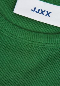 Grønn rillet stoff med rund hals; har et hvitt merke med blå tekst "JJXX." Teksturen er jevn, noe som fremhever stoffets elastisitet.