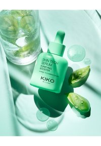 Botella de suero verde menta con cuentagotas; etiquetada "KIKO MILANO SKIN TECH SERUM." Alrededor hay gotas y hojas de albahaca sobre una superficie verde suave.