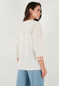 Top beige chiaro con maniche a tre quarti, caratterizzato da un orlo testurizzato con dettagli a frangia. Indossato con pantaloni larghi blu.