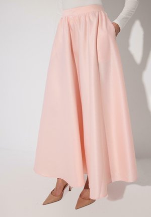 Ženska v dolgi, tekoči, svetlo roza krilu in golih, špičastih petah, stoji ob preprosti beli steni.