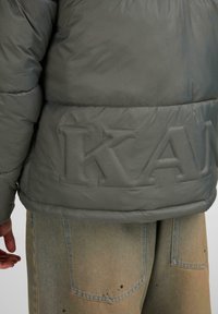 Hall mõõduga hallist polsterdatud jakk, mille hemist paistab silma väljapaistvalt reljeefne "KA" logo. Kombineeritud heledate teksadega, millel on tuhm tekstuur ja nähtavad taskud.
