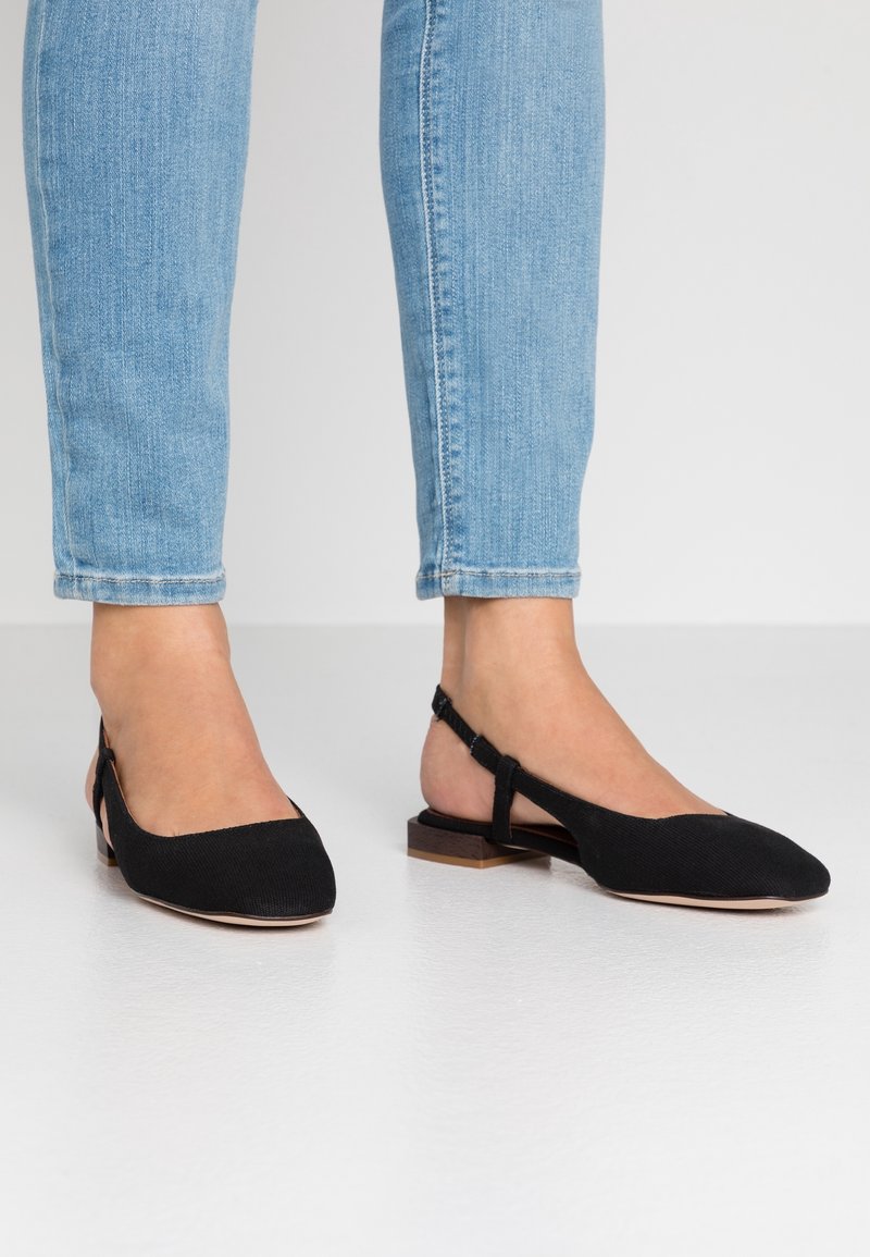 Ballerines noires à bride arrière avec un bout pointu, dessus en tissu, petit talon carré et détail découpé sur les côtés. Portées avec un jean bleu clair.