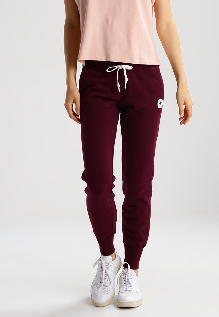Bordeaux sweatpants met elastische boorden en een trekkoord in de taille, met een witte logopatch op de linker bovenbeen, gecombineerd met witte sneakers.