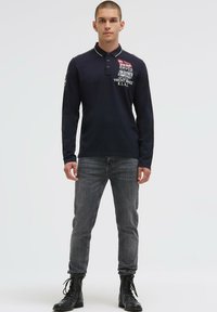 Junger Mann, der ein marineblaues Langarm-Poloshirt mit weißen Textgrafiken, graue Jeans und schwarze Schnürstiefel trägt und vor einem einfachen Hintergrund steht.