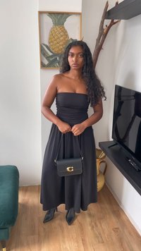 Abito nero senza spalline con corpetto arricciato, gonna lunga fino al pavimento, abbinato a tacchi neri a punta e una piccola borsa nera con logo dorato.