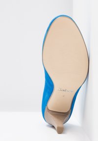 Chaussure à talon haut en daim bleu reposant la semelle vers le haut contre un coin blanc, montrant la semelle beige avec la marque "Gabor" et la taille 4 indiquée.