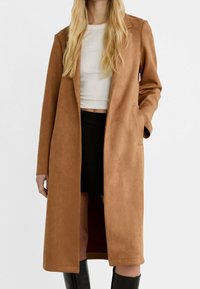 Mujer con un abrigo largo color camel sobre una camiseta blanca corta, falda negra y botas negras hasta la rodilla, de pie con una mano en el bolsillo.