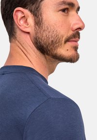 Close-up van het zijaanzicht van een man met kort donker haar, een getrimde baard en een blauwe crewneck shirt tegen een effen lichte achtergrond.