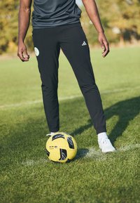 Zwarte sportbroek met taps toelopende benen en een Adidas-logo, gecombineerd met witte sneakers, geplaatst naast een gele voetbal op gras.