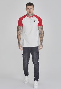Cămașă albă cu mâneci scurte și mâneci raglan roșii, având un logo negru. Asortată cu pantaloni cargo negri și adidași albi. Modelul stă în centru.