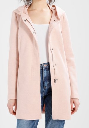 Cappotto cappuccio rosa chiaro con chiusura a snap e zip, maniche lunghe, tessuto testurizzato e tasche frontali, indossato sopra una maglietta bianca e jeans blu.