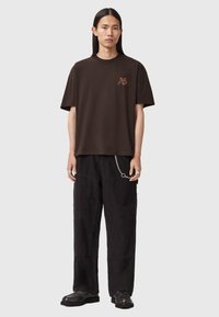 Brązowy bawełniany t-shirt z małym logo na piersi, zestawiony z czarnymi sztruksowymi spodniami z dużymi kieszeniami i ozdobą w formie łańcucha.